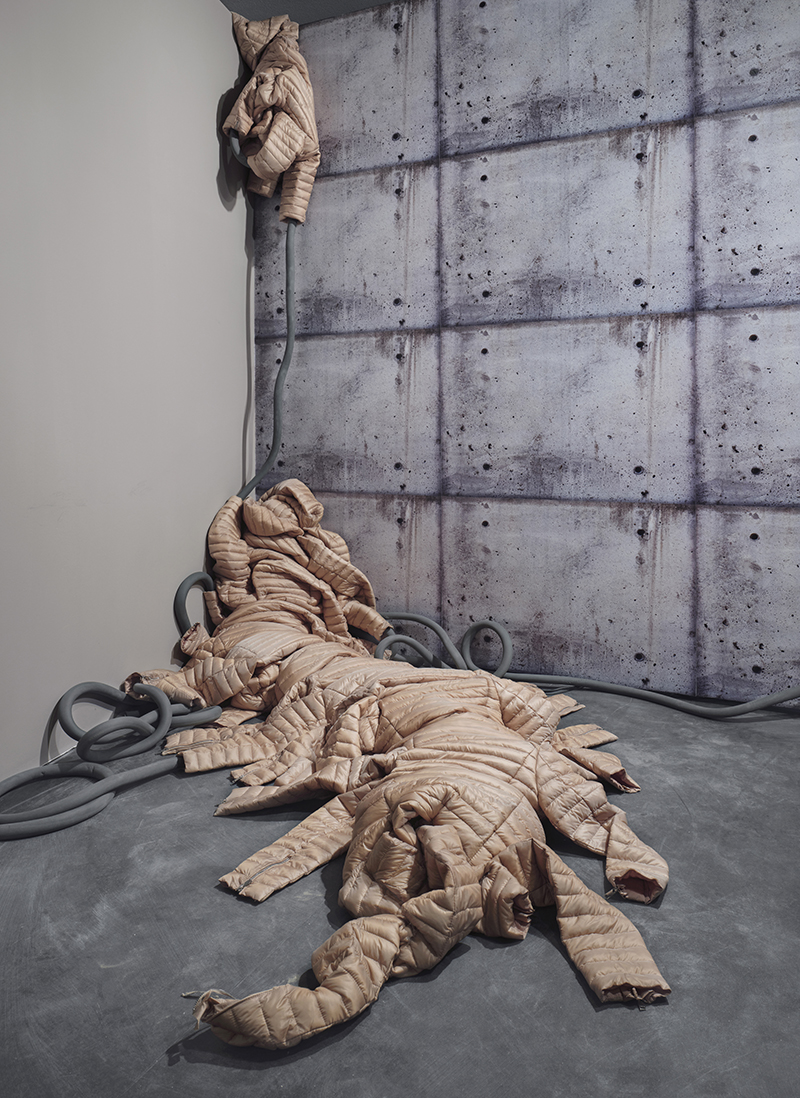 A creature sewn from puffy jackets against fake concrete wallpaper