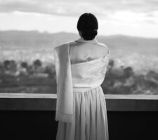 black & white photograph of a woman seen from behind wearing an elegant dress, standing at a balustrade infront of a wide landscape
