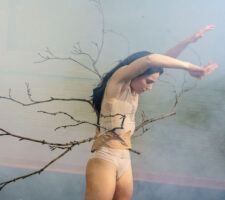 Photograph of a performer dancing in a foggy studio with her arms in the air and surrounded by tree branches that are stick in her belt