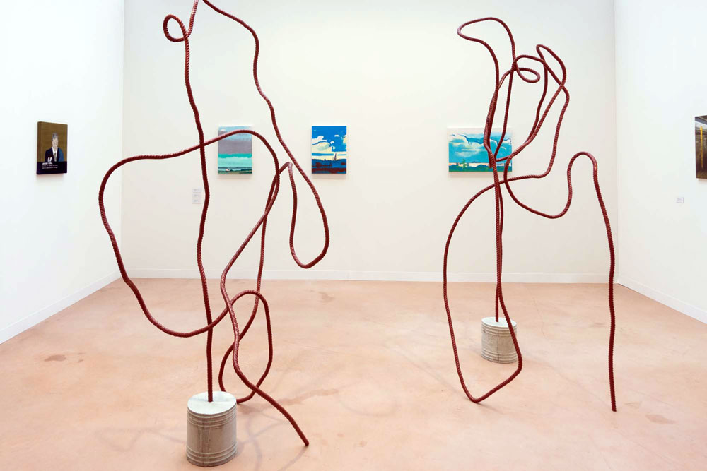 Two red Sculptures made of rebar standing in a fair booth with three blue paintings hanging on the back wall