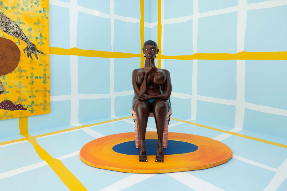 Sculpture of a Woman looking into the camera, sitting on a chair in an art fair booth with bright blue painted walls and yellow and white geometrical lines 